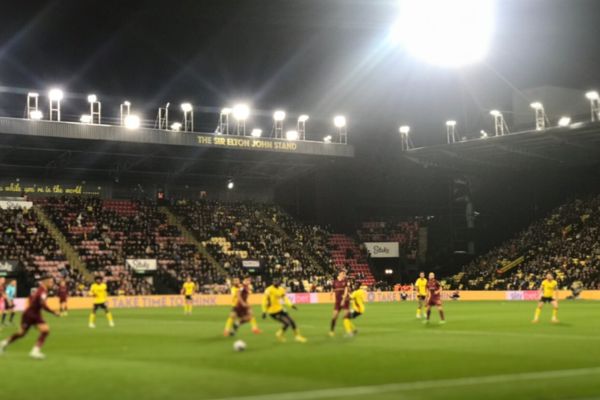 Sir Elton John Stand at Vicarage Road stadium during a match, viewed from the opposite pitch side under floodlights, with the words 'Sir Elton John Stand' visible on the roof.