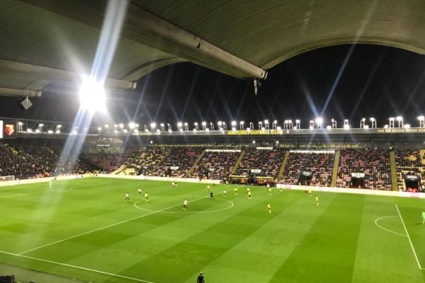 View of the Sir Elton John Stand at Vicarage Road at night, illuminated by floodlights, as seen from the Graham Taylor Stand.