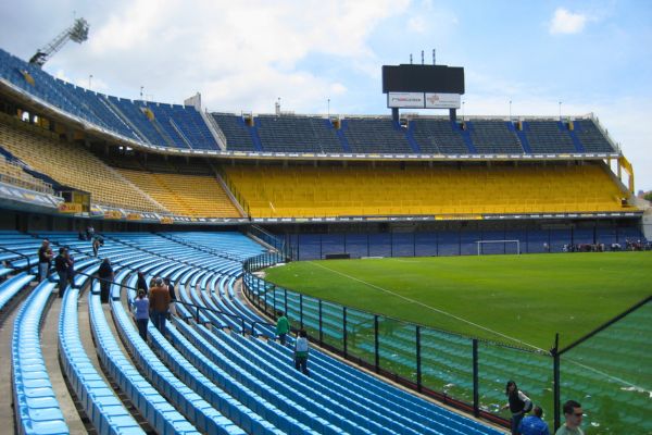 Tourists walking through the seating sections of La Bombonera stadium, surrounded by rows of blue and yellow seats with a view of the pitch below.