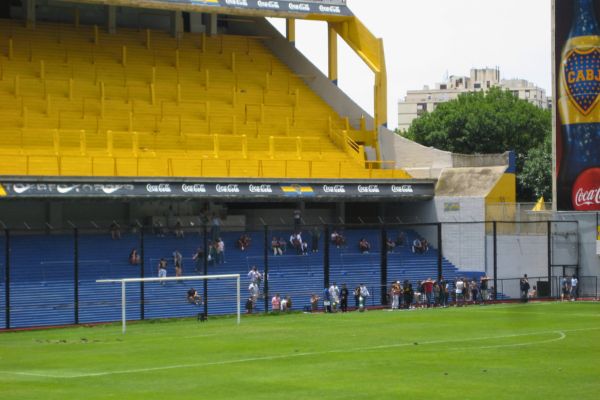 A tour group in the distance walks along the terraces behind the goal at La Bombonera stadium, with steep blue and yellow stands towering around them.