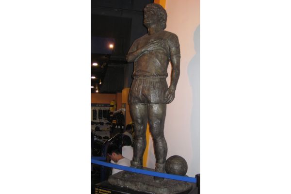 A bronze statue of Diego Armando Maradona in the Boca Juniors Museum, depicting the legendary footballer in a triumphant pose, wearing Boca Juniors' iconic kit.