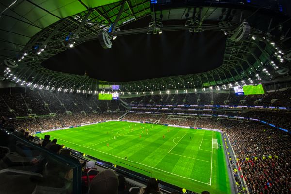 A vibrant scene inside Tottenham Hotspur Stadium during an evening football match, with bright floodlights illuminating the pitch and a sea of fans in the stands.