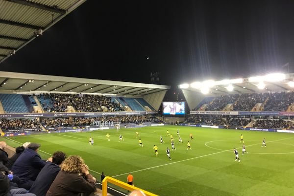 A thrilling football match at The Den, home of Millwall FC, illuminated by floodlights.