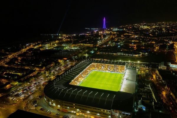 Aerial view of Bloomfield Road stadium lit up at night, showcasing its vibrant lights and surrounding Blackpool cityscape.