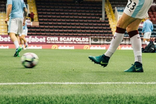 A player kicking the ball during a match on the Turf Moor pitch, with the stands in the background.