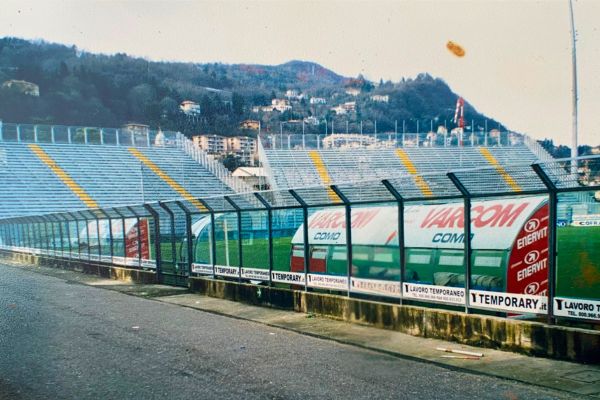 Empty Como Stadium from pitch level behind the dugouts, showcasing the stands and field.