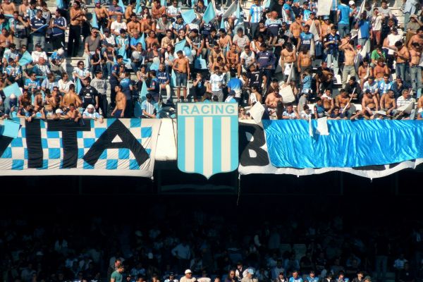 Racing Club fans in the stands with a large club badge displayed on the upper balcony of Estadio Presidente Perón.