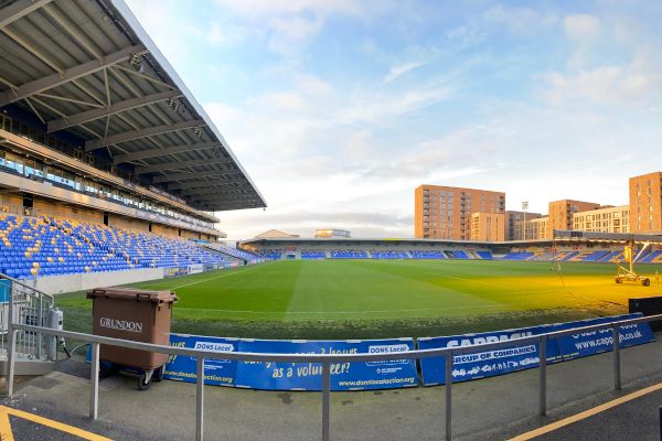 An inside view of the empty Plough Lane Stadium, showcasing the modern seating, pitch, and surrounding stands under a blue sky.