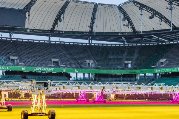 Empty stands at Volkswagen Arena in Wolfsburg with an artificial lighting rig illuminating the pitch for grass maintenance.