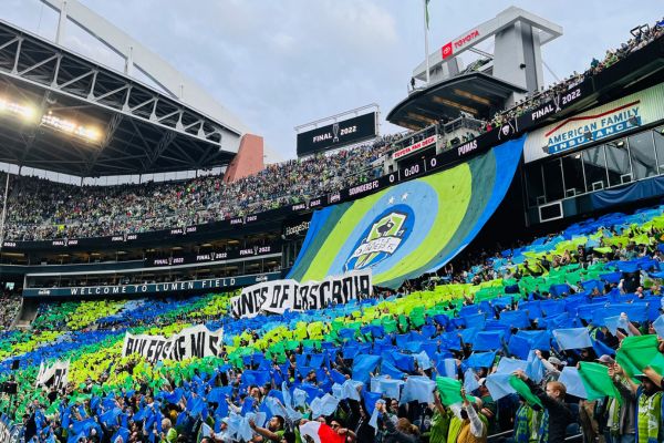 Packed stands of Seattle Sounders fans at Lumen Field on matchday, waving flags and cheering in vibrant green and blue team colors.