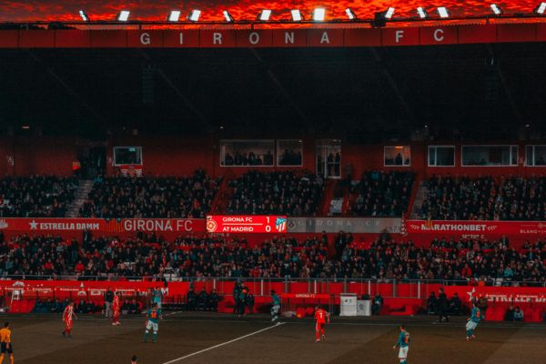 A football match in progress at Estadi Montilivi, the home of Girona FC, with players on the field and fans cheering from the stands.