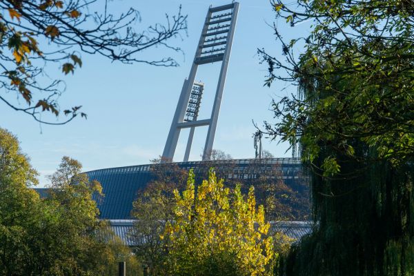 A modern stadium exterior during the day, with sleek architecture, clear blue skies, and people walking nearby.