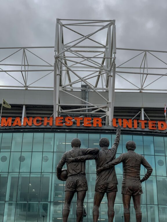 The "United Trinity" statue in front of Old Trafford, with the red "Manchester United" sign visible above.